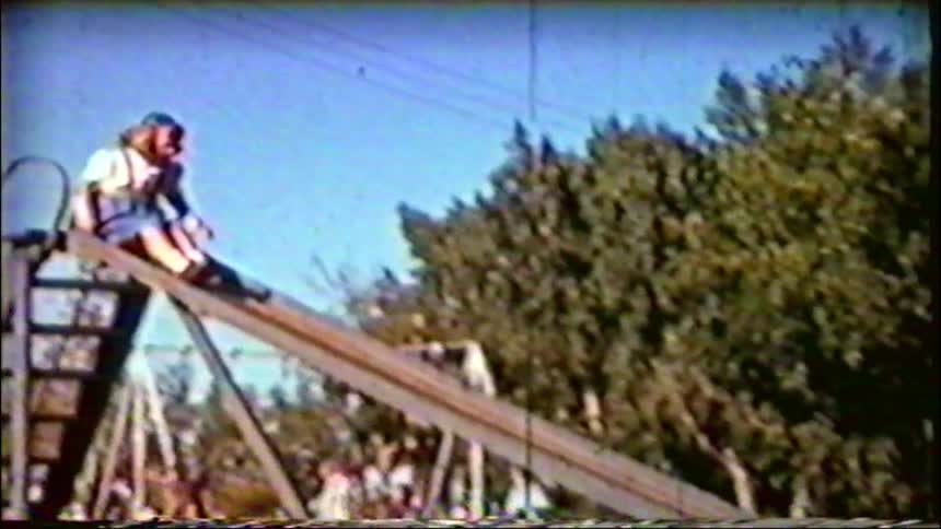 Vintage home movie footage shows a happy young girl sliding down a metal slide at a park on a sunny day