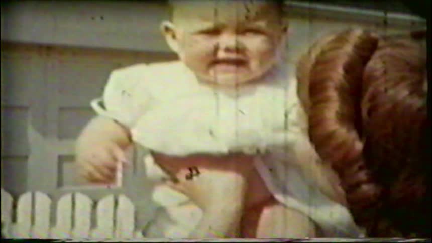 A mother with glasses smiles and holds her fussy but curious baby outdoors in a vintage home movie clip