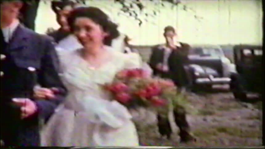 A smiling bride and her groom in a military uniform walk arm-in-arm with their wedding party in charming vintage footage