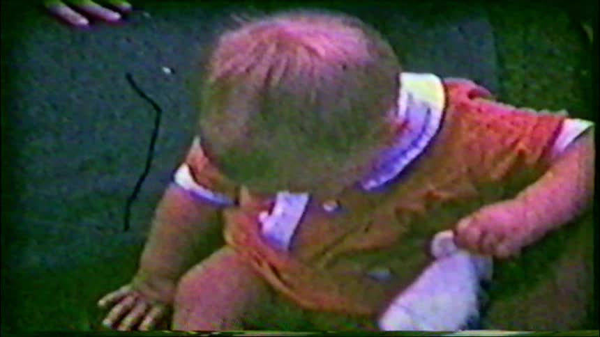 A grainy, vintage home movie captures a close-up of a baby in a red romper sitting on the green grass