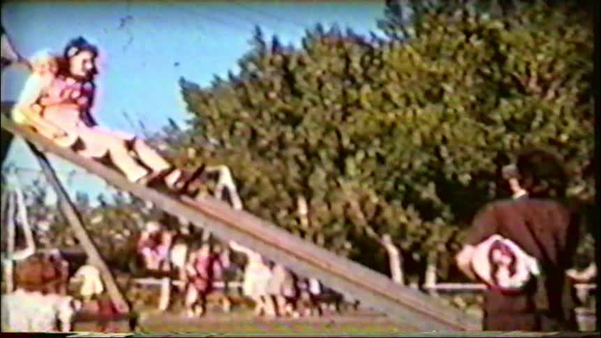 A vintage home movie captures a young girl in a pink dress sliding down a metal slide at a sunny park