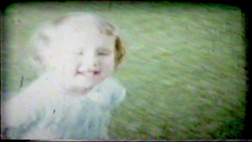 An overexposed, vintage home movie shows a smiling baby girl sitting on a grassy lawn