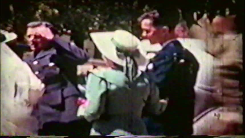 A vintage 1940s home movie shows a groom in a military uniform helping his bride into a car as wedding guests throw confetti