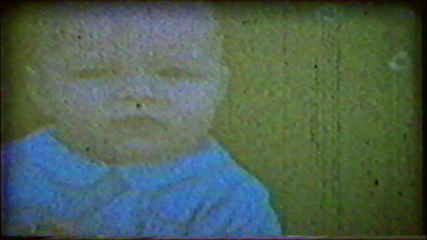 A grainy, vintage home movie shows a close-up of a sad-looking baby in a blue shirt