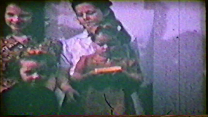 A grainy, vintage home movie captures a family with several women and young children posing together for a group portrait