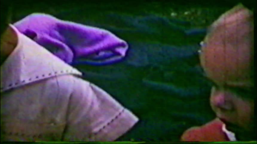 A close-up of a young child's face reveals their older sibling behind them in grainy vintage home movie footage