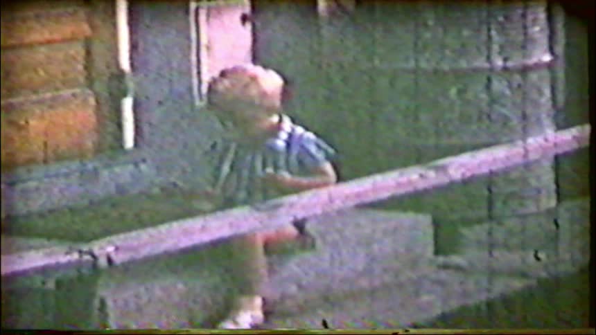 A small child sits alone on some steps in a grainy, nostalgic home movie clip from the past