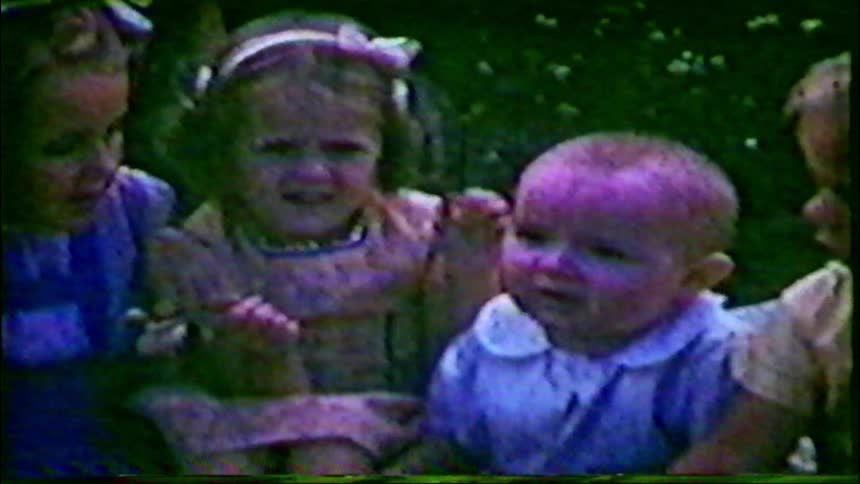 A grainy vintage home movie captures a family with young children and a baby posing together outdoors