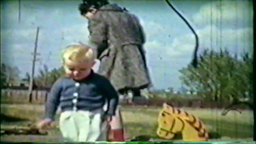 Vintage home movie captures a toddler boy playing in a dirt yard with his rocking horse toy