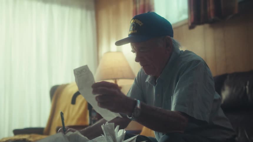 An elderly Air Force veteran with a concerned expression sits in his living room and carefully reviews his bills and receipts