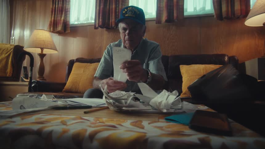 An elderly veteran wearing an Air Force hat sits on his couch and carefully reviews his bills and receipts while managing his finances