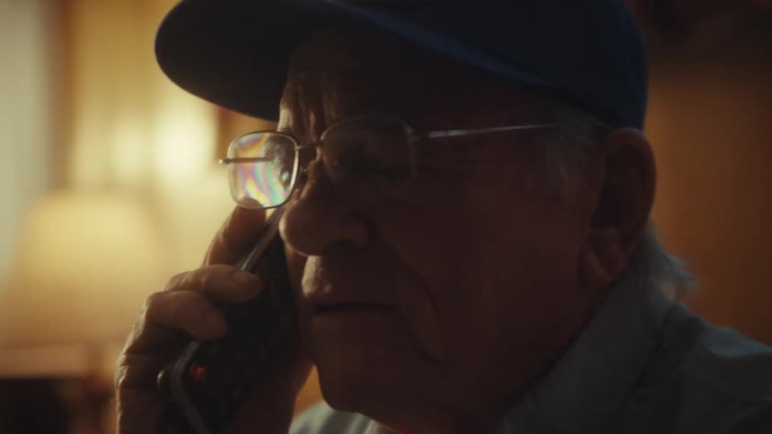 A concerned elderly Air Force veteran wearing glasses and a hat talks on the phone in his home