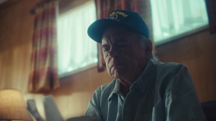 A pensive elderly veteran wearing a U.S. Air Force hat sits alone in his home and solemnly reads a letter