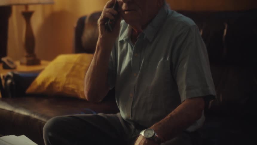 An elderly veteran with a concerned expression talks on the phone while sitting in his living room