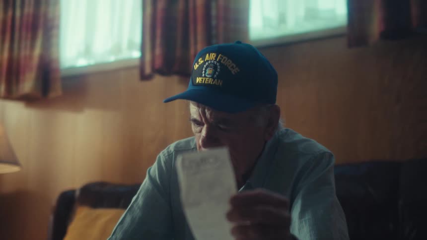 An elderly US Air Force veteran sits with a pensive expression as he carefully goes over his bills in his living room