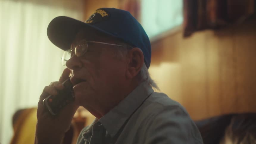 An elderly Air Force veteran listens intently to a serious phone call while sitting on his couch at home