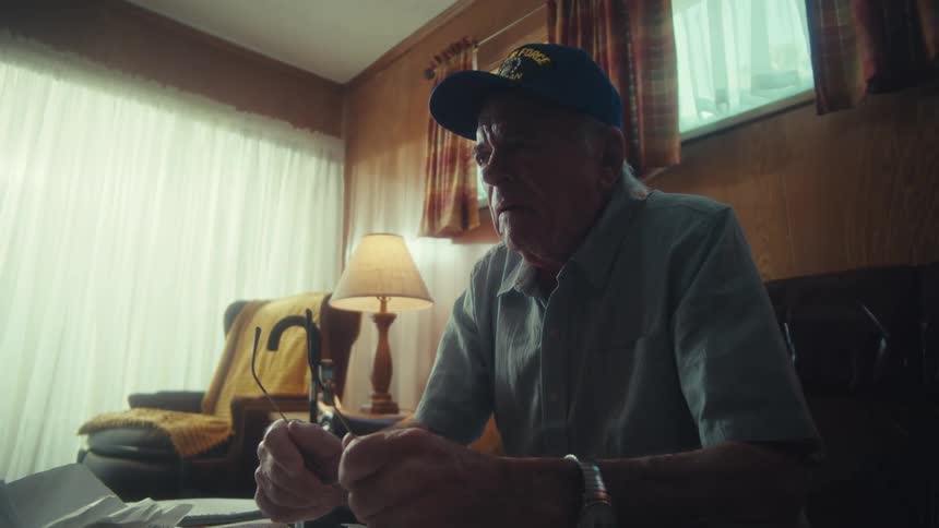 Elderly Air Force veteran sits pensively in his living room while cleaning his eyeglasses