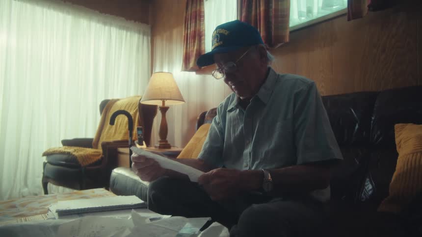 An elderly veteran sits on a leather couch in his home, diligently sorting through a pile of papers and bills