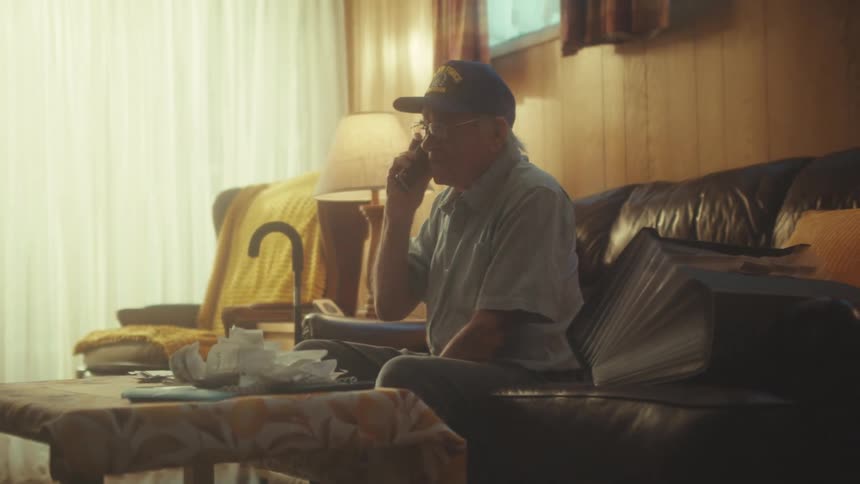 An elderly man in a veteran's cap sits on a leather couch in his quiet living room, having a conversation on the phone as the camera slowly pans