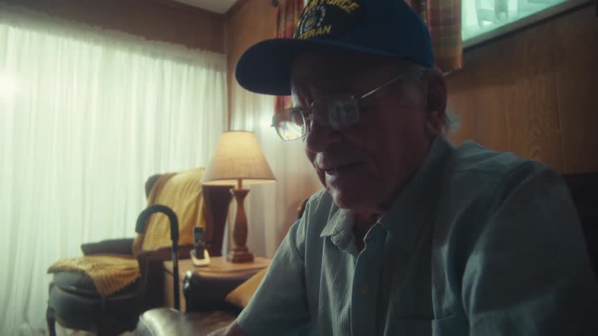 An elderly Air Force veteran in glasses and a cap sits in his sunlit living room and thoughtfully writes on a piece of paper