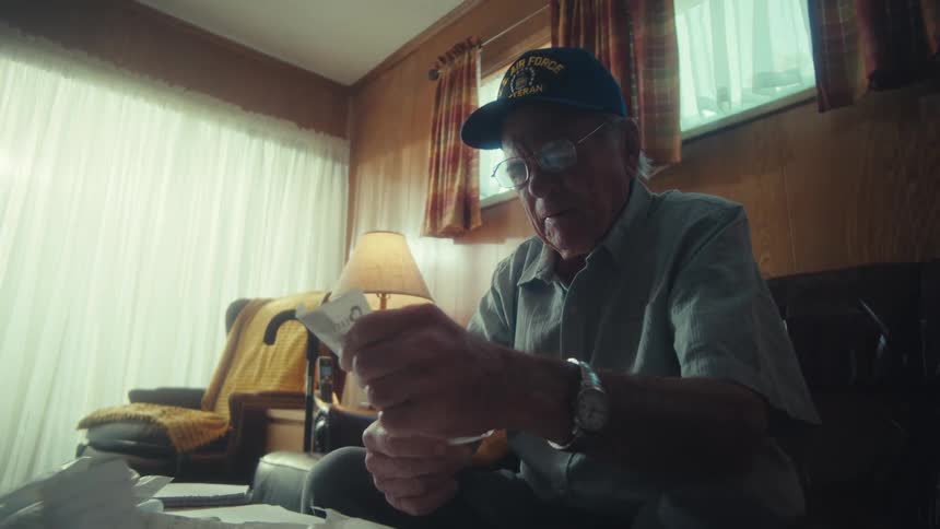An elderly Air Force veteran sits in his dimly lit living room, carefully sorting through his bills and receipts