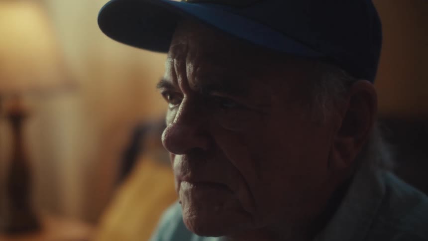 Close up of an elderly U.S. Air Force veteran looking worried and stressed as he sits in a dimly lit room with a pile of receipts in the foreground
