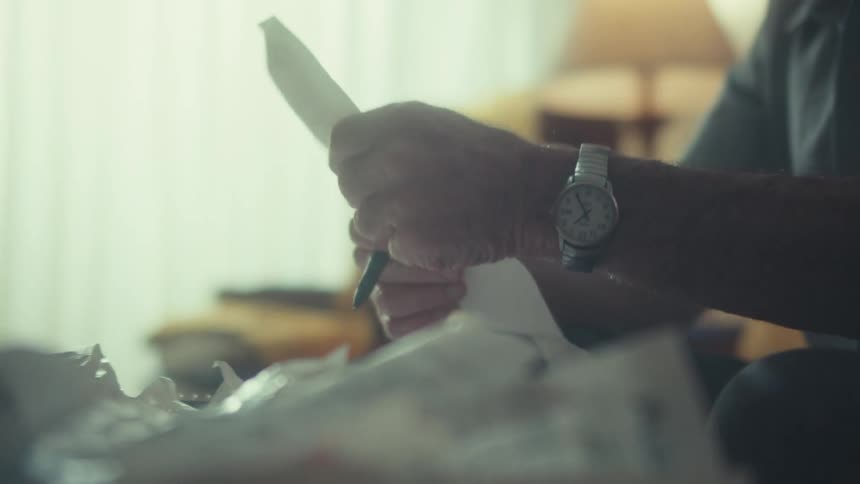 An elderly man meticulously reviews a long receipt while managing his finances at a table cluttered with bills