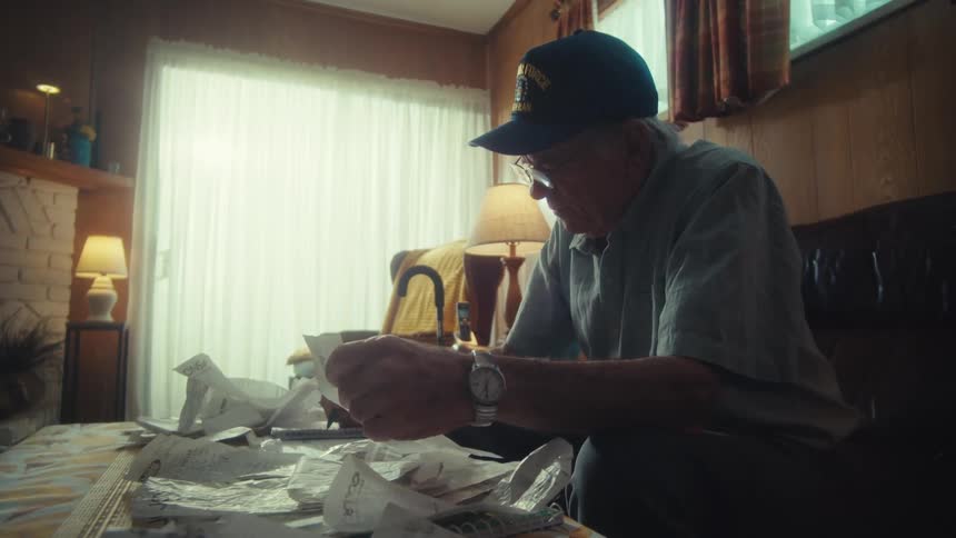 An elderly Air Force veteran wearing a cap sits at his coffee table meticulously sorting through bills and receipts