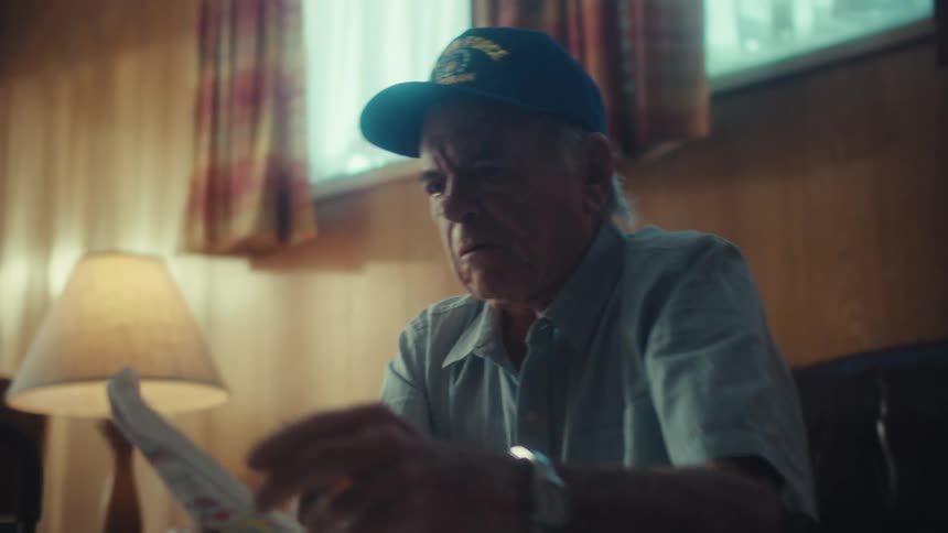 A worried elderly veteran sits in his dimly lit home, anxiously sorting through a large pile of bills and receipts