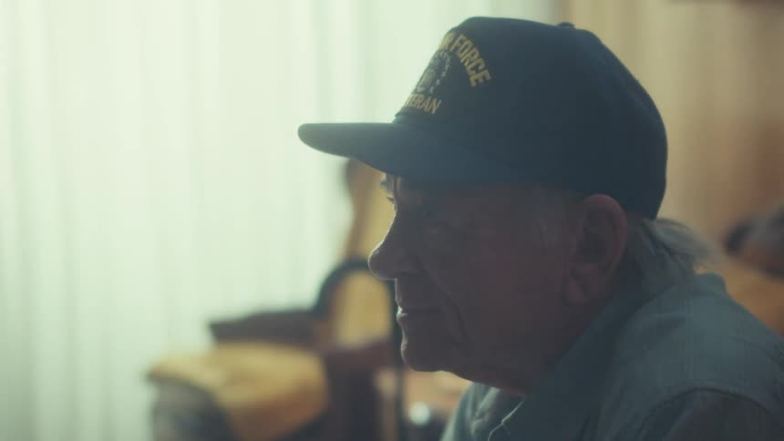 A thoughtful elderly Air Force veteran wearing a veteran's cap sits quietly at home, bathed in soft window light