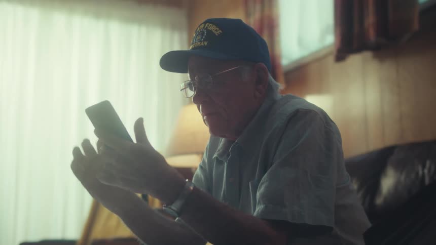 A thoughtful elderly Air Force veteran sits in a softly lit living room looking at his smartphone