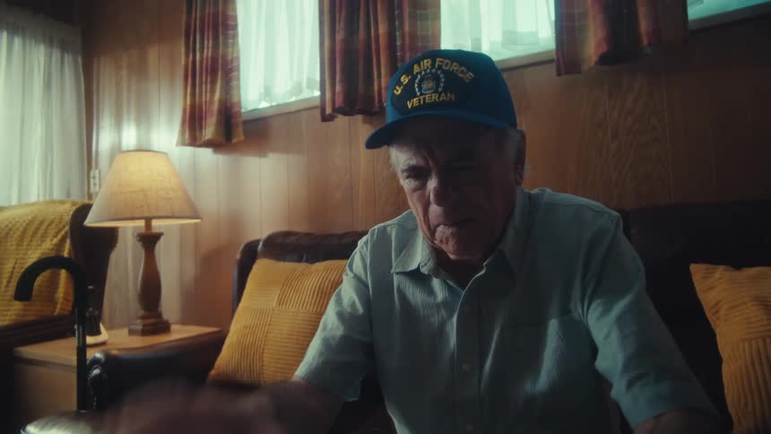 An elderly Air Force veteran sits on his couch looking overwhelmed and stressed as he sorts through a large pile of bills and receipts