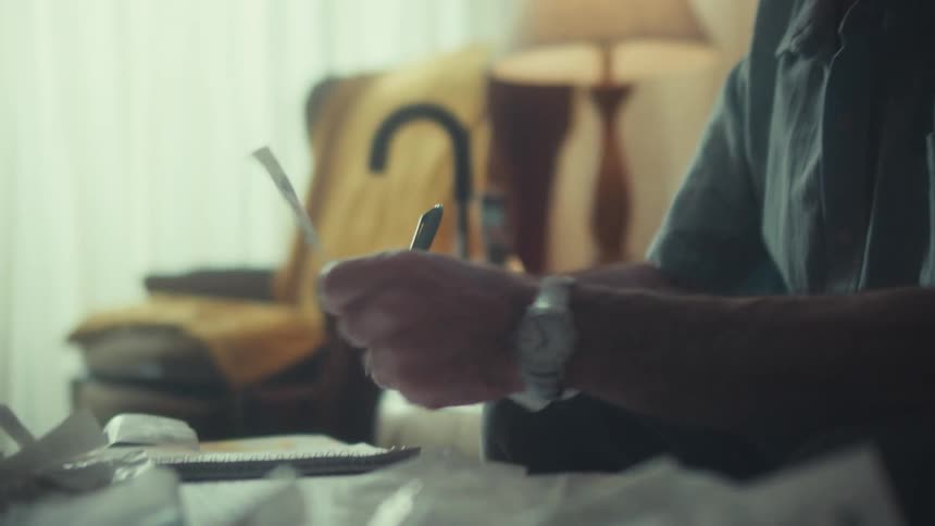 An elderly Air Force veteran meticulously sorts through receipts and writes in a notebook while managing his personal finances at home