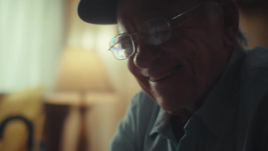 A cinematic close-up of a smiling elderly man with glasses and a cap using a smartphone in a warmly lit room