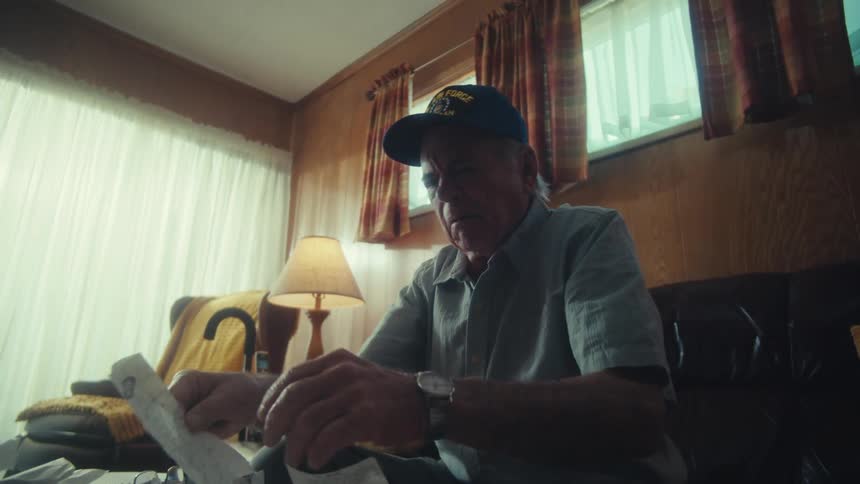 An elderly veteran becomes overwhelmed and frustrated while sorting through his finances in his dimly lit living room