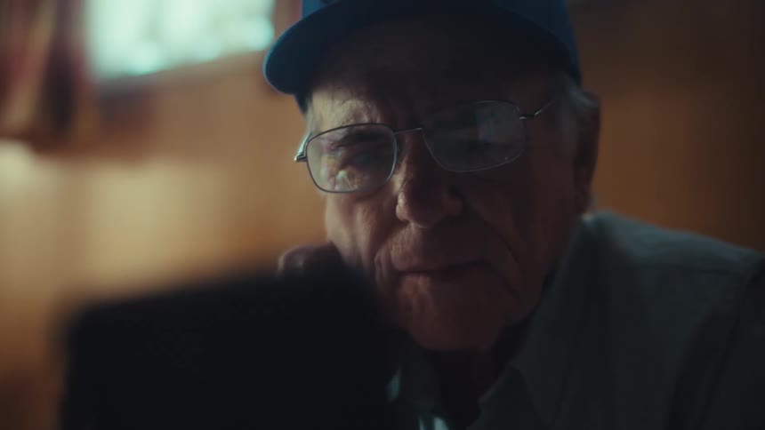 A thoughtful elderly U.S. Air Force veteran wearing a cap and glasses watches something on his smartphone in his home