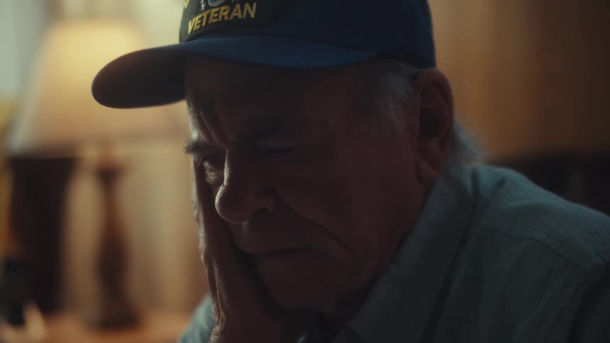 A solemn elderly Air Force veteran sits alone in a dimly lit room, reflecting on painful memories