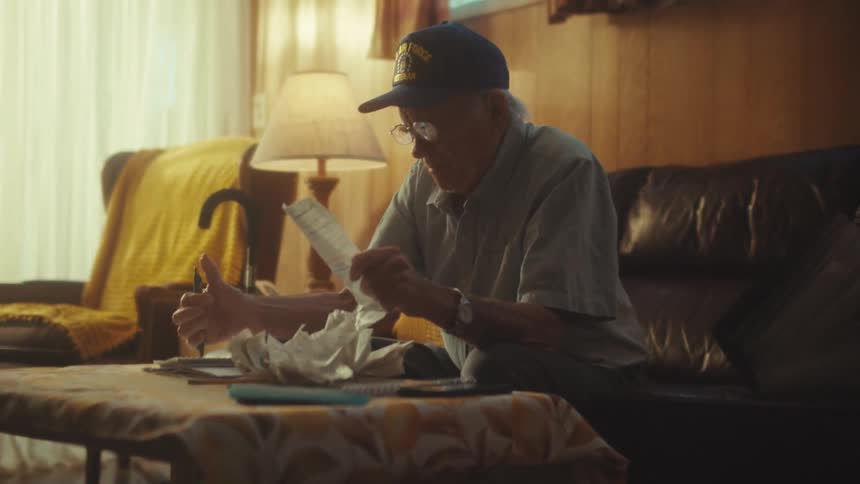 An elderly Air Force veteran carefully reviews his bills and finances while sitting on the couch in his sunlit living room