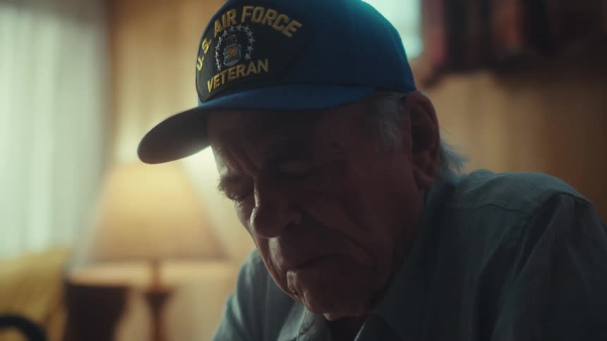 A close-up shot of a distressed elderly veteran sitting alone in his living room, appearing worried and overwhelmed while looking at papers