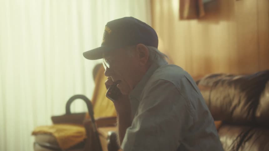 An elderly Air Force veteran wearing a baseball cap sits on his couch and talks on a cordless phone at home