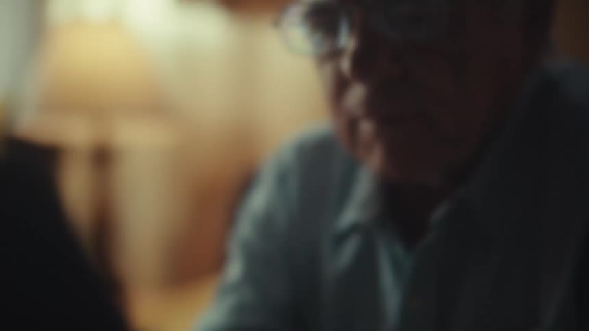 A cinematic close-up of a thoughtful elderly man in a hat and glasses as he scrolls on his smartphone in a dimly lit room