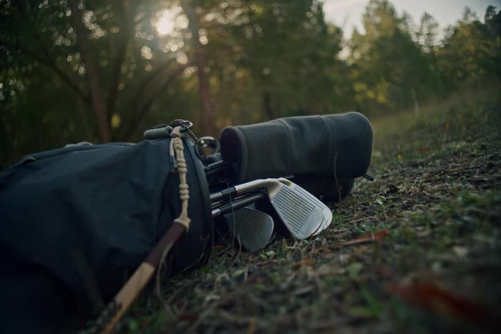A close-up of a golf bag with clubs being thrown down on the grass next to another bag in a sun-drenched forest