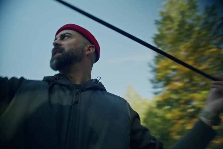 A bearded man wearing a red beanie assembles a long tent pole while setting up camp in a sunny, wooded area