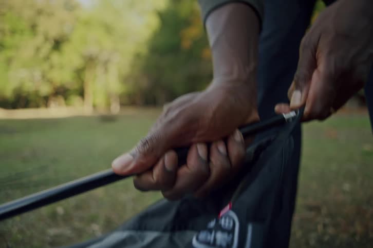 A close-up shot shows a man's hands carefully threading a flexible pole through the fabric sleeve while setting up a tent in a grassy park