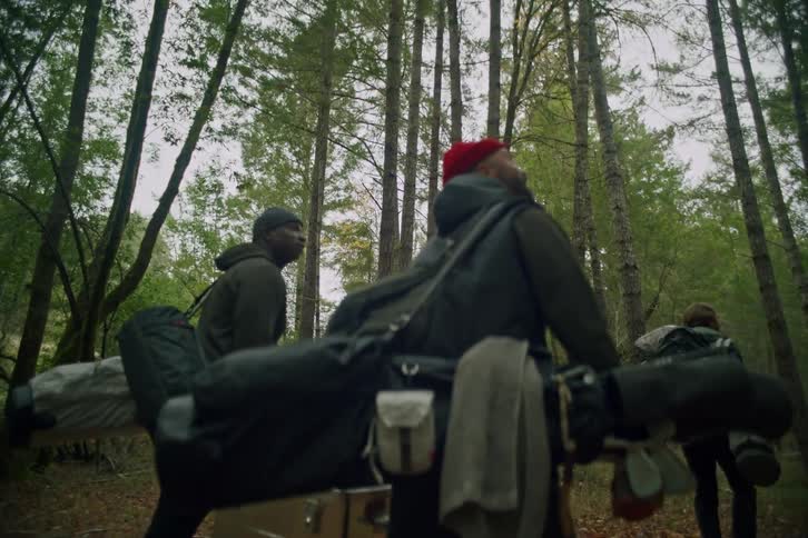 A low-angle follow shot of a group of men carrying golf bags and other gear as they hike through a dense forest