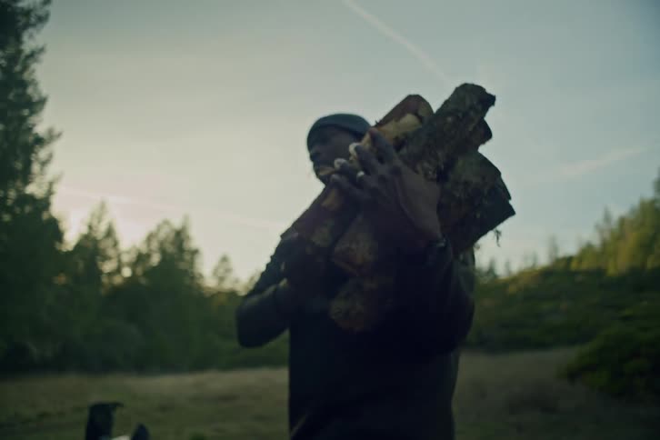 Slow motion shot of a man carrying a bundle of firewood through a campsite in a forest clearing at dawn