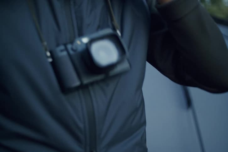 A bearded photographer in a red beanie and vest adjusts his clothes at a campsite in the woods