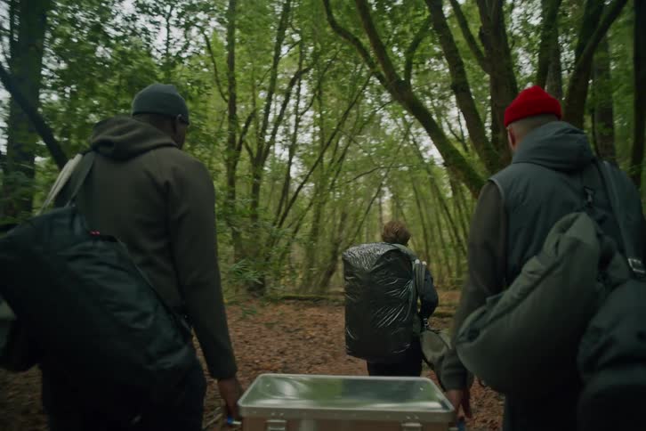 A team of three carries heavy equipment while hiking together through a dense, lush forest