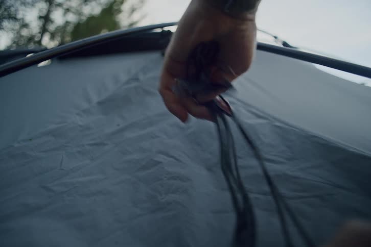 A person's hands untangle and tighten the guy line on top of a grey tent while setting up a campsite