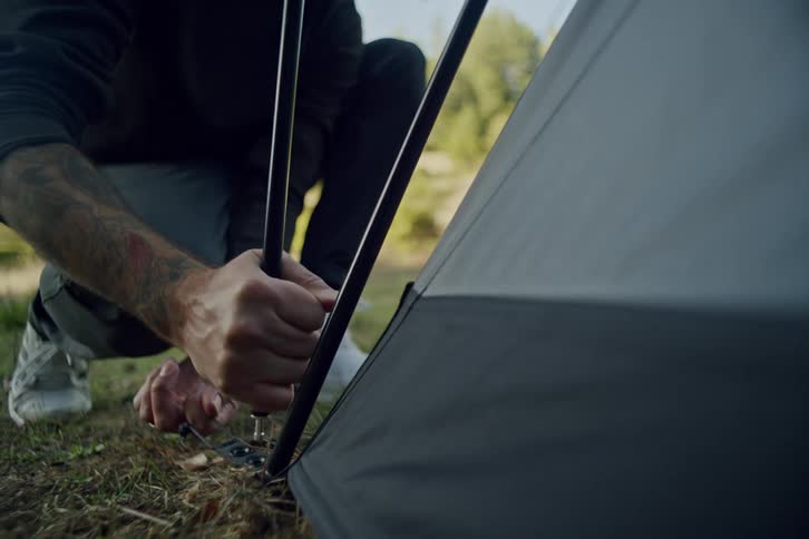 A man in a red beanie carefully assembles the poles of a tent in a sunlit forest clearing in slow motion
